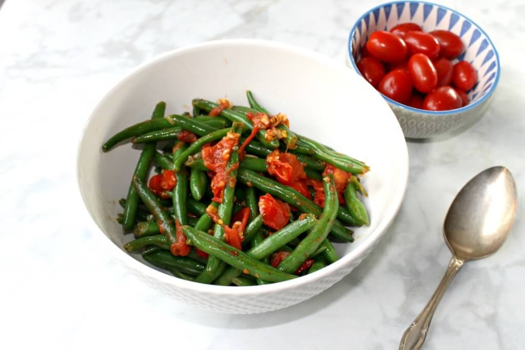 overhead shot of green beans with tomatoes and garlic, with a spoon and a small bowl full of cherry tomatoes on a white marble countertop
