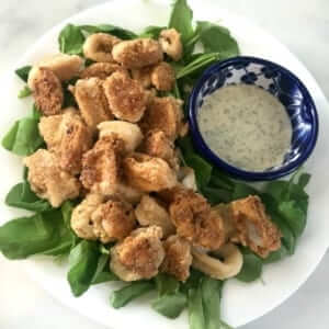 bowl of fried calamari on a white plate with greens and a small bowl of sauce