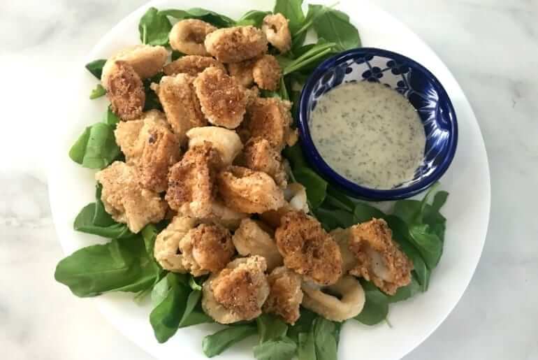 bowl of fried calamari on a white plate with greens and a small bowl of sauce