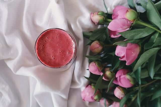 a pink smoothie next to a bunch of pink peonies on a ruffled bedsheet