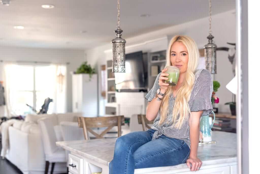 a blond woman sipping an anti-stretch mark smoothie with collagen powder sitting on her kitchen counter 