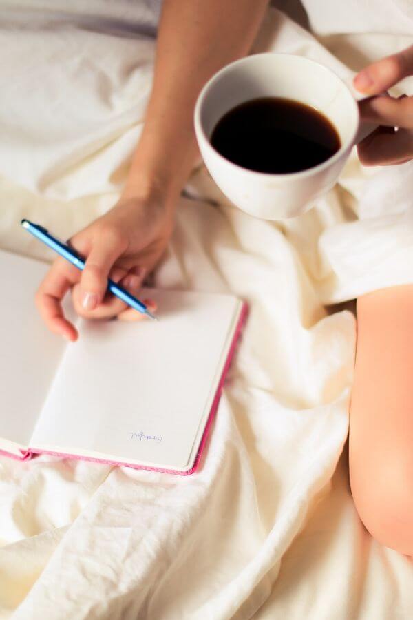 a woman writing a to-do list in bed with a cup of coffee in hand 