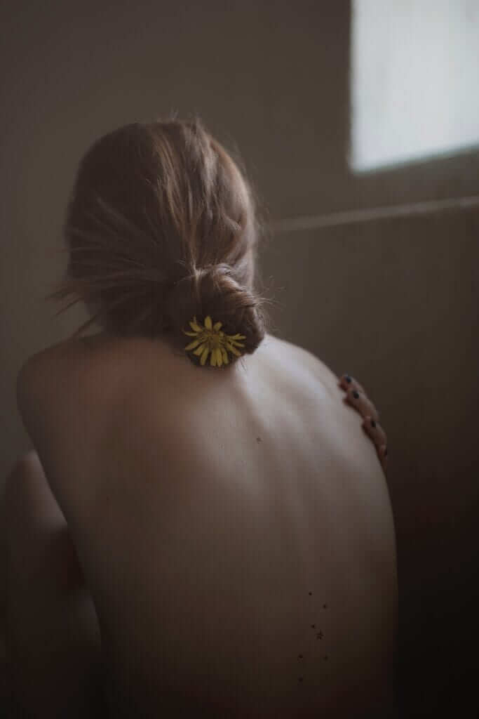 a woman with a flower in her hair sitting with her back towards the camera hugging her naked breasts to hide them from sight