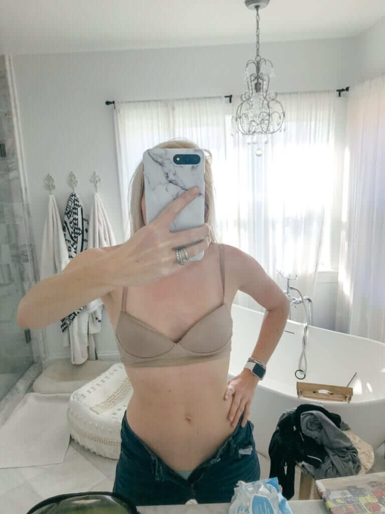 a woman in a beige bra and dark jeans taking a selfie in front of the mirror